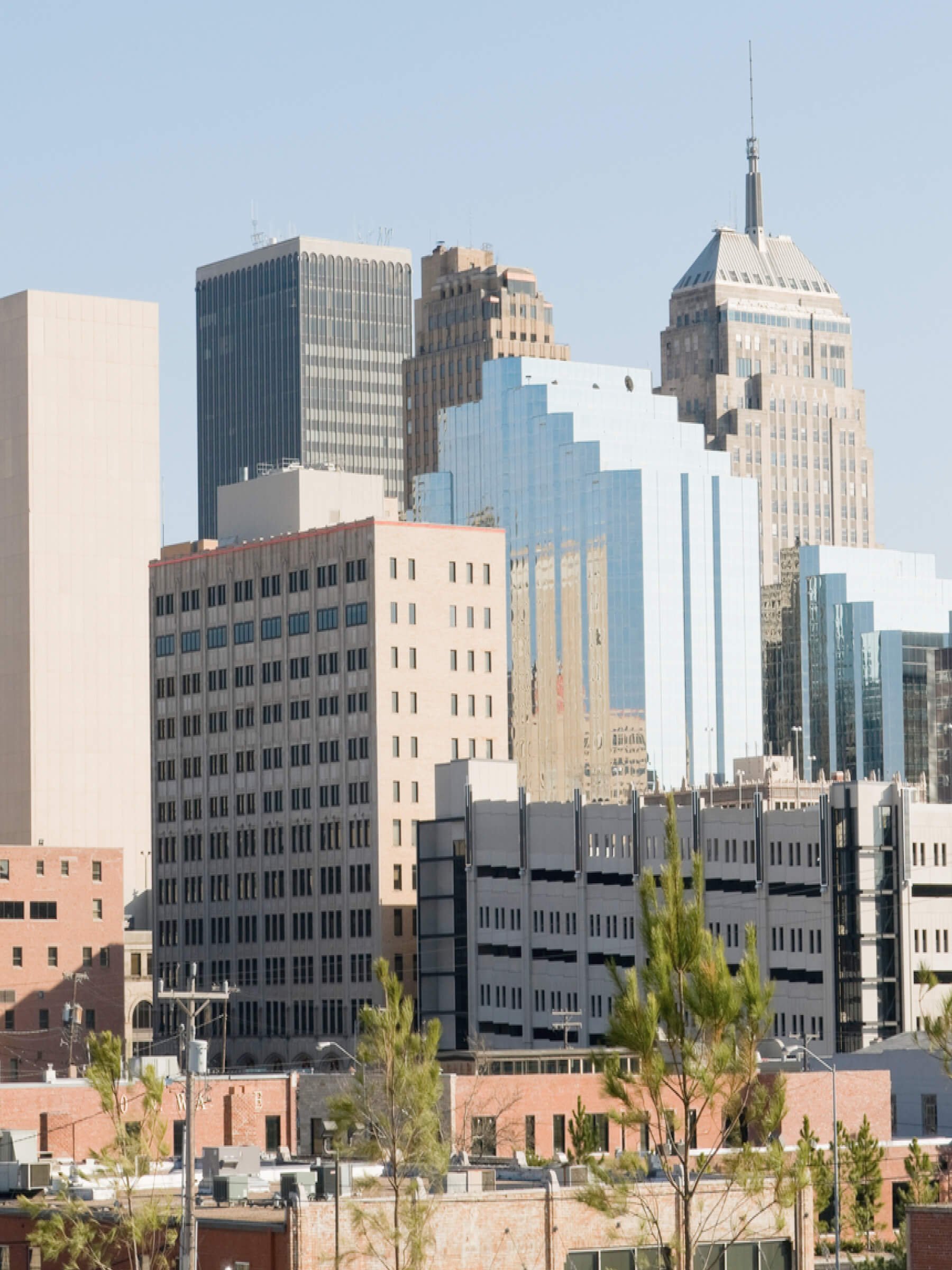 City skyline with various tall buildings, some with reflective glass windows, and green trees in the foreground.