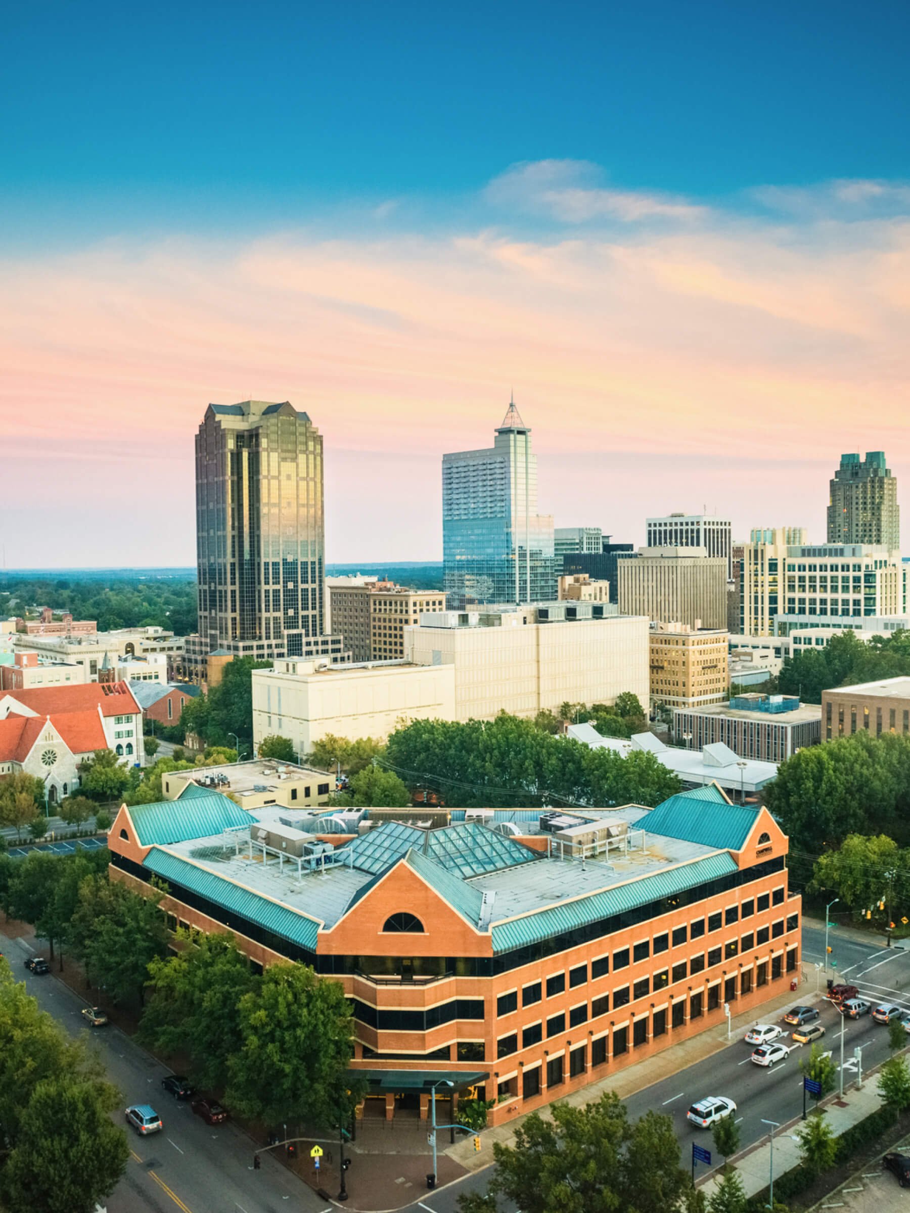 A city skyline during sunset with modern high-rise buildings, a mix of older and newer architecture, and green trees in the foreground.
