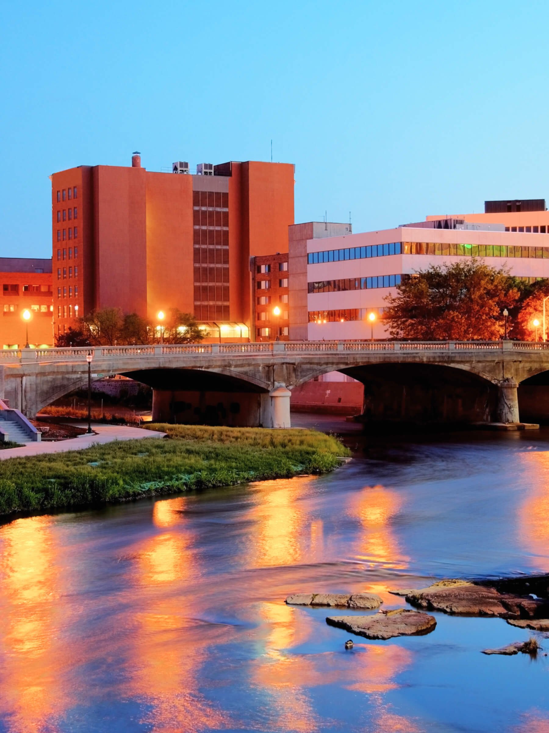 Cityscape of Sioux Falls South Dakota at dusk with buildings, a bridge over a river, and reflections of lights on the water.