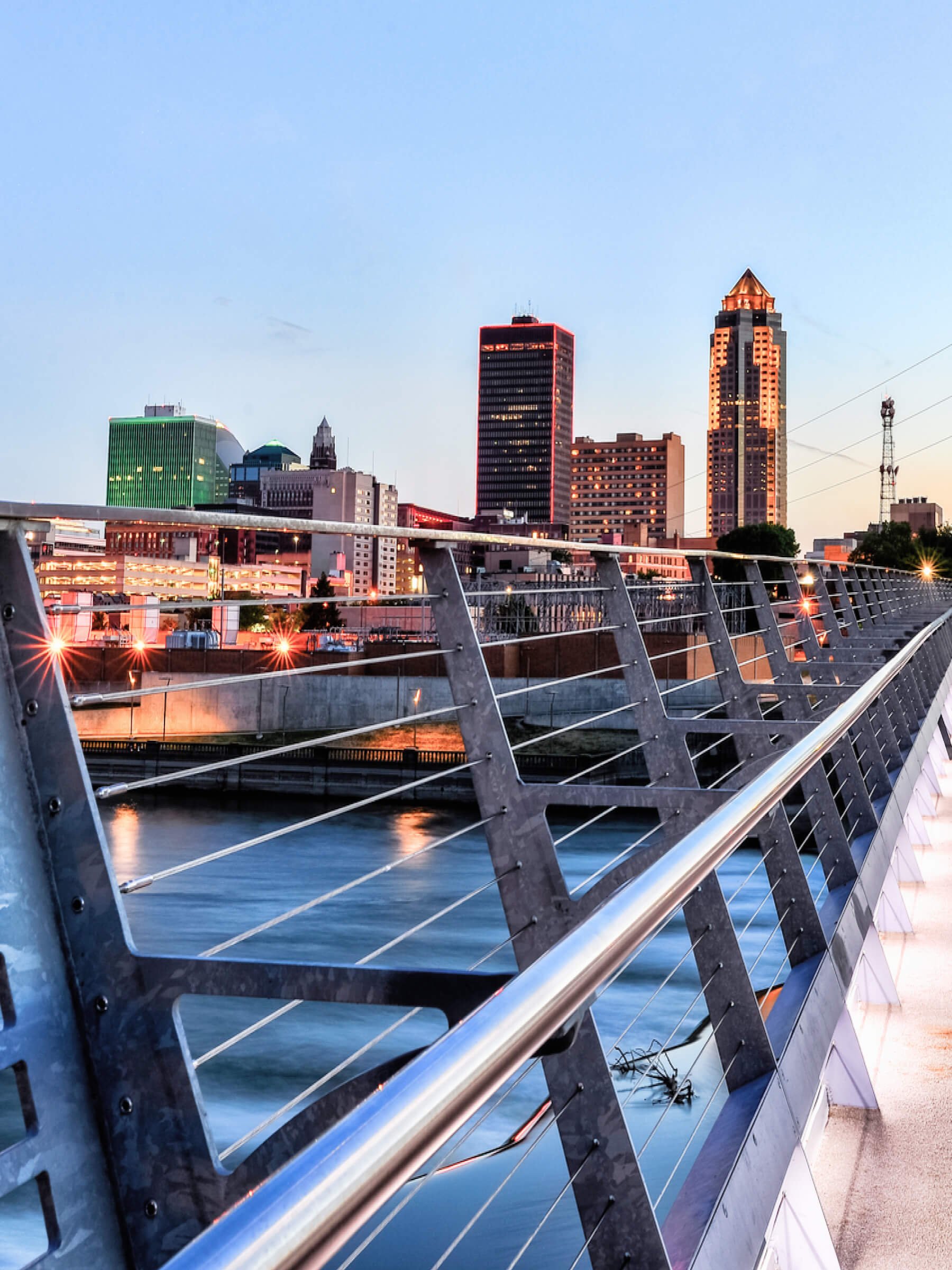 City skyline at sunset with buildings, a bridge railing in the foreground, and a river below.