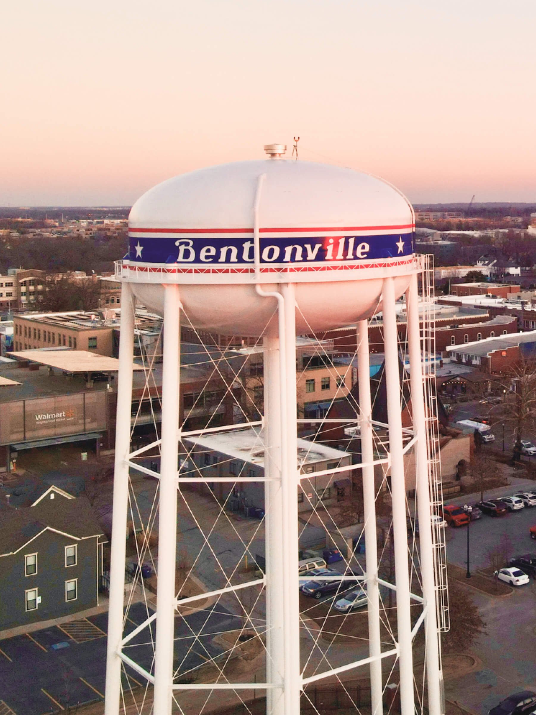 Water tower with 'Bentonville' written on it, surrounded by buildings, parking lots, and streets at dusk