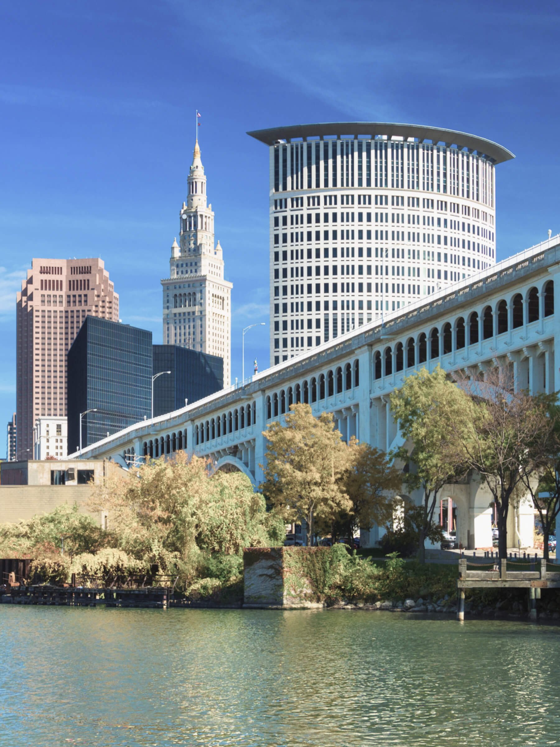 City skyline with a river in the foreground, featuring various tall buildings, including a historic clock tower and modern high-rises, under a clear blue sky.