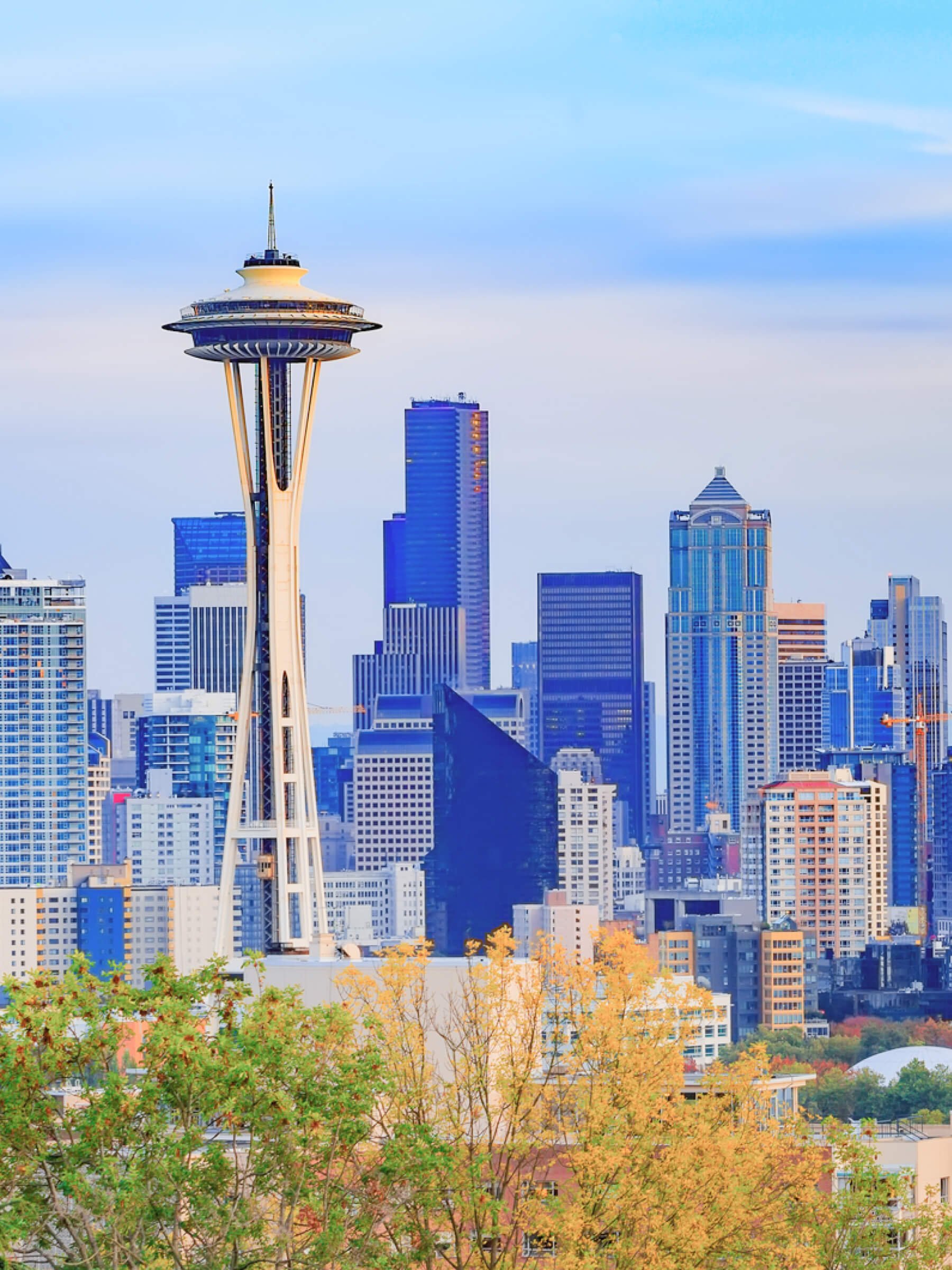 Seattle skyline featuring the Space Needle with tall modern buildings and trees with autumn leaves in the foreground.