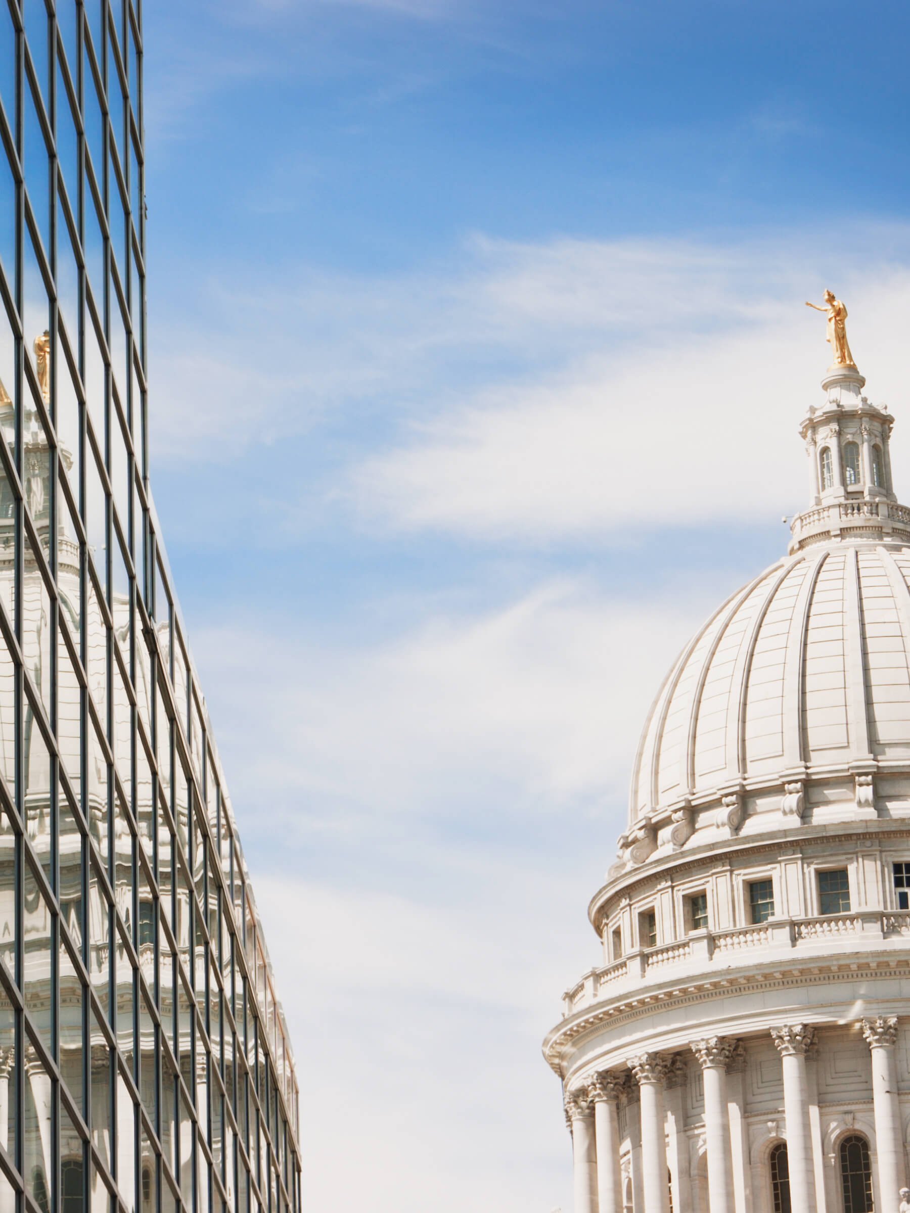 The image shows the U.S. Capitol building with a prominent dome topped with a statue, and a modern glass building reflecting the Capitol on the left side, under a partly cloudy blue sky.
