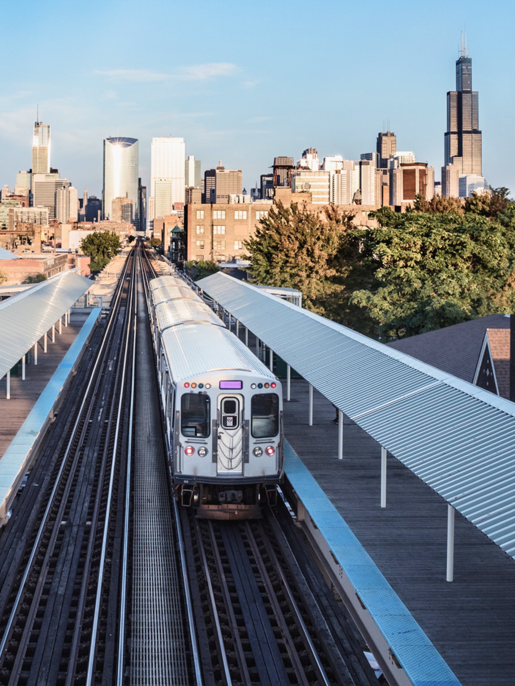 An elevated train station with a silver train approaching, set against a city skyline with tall buildings and skyscrapers in the background, under a clear blue sky.