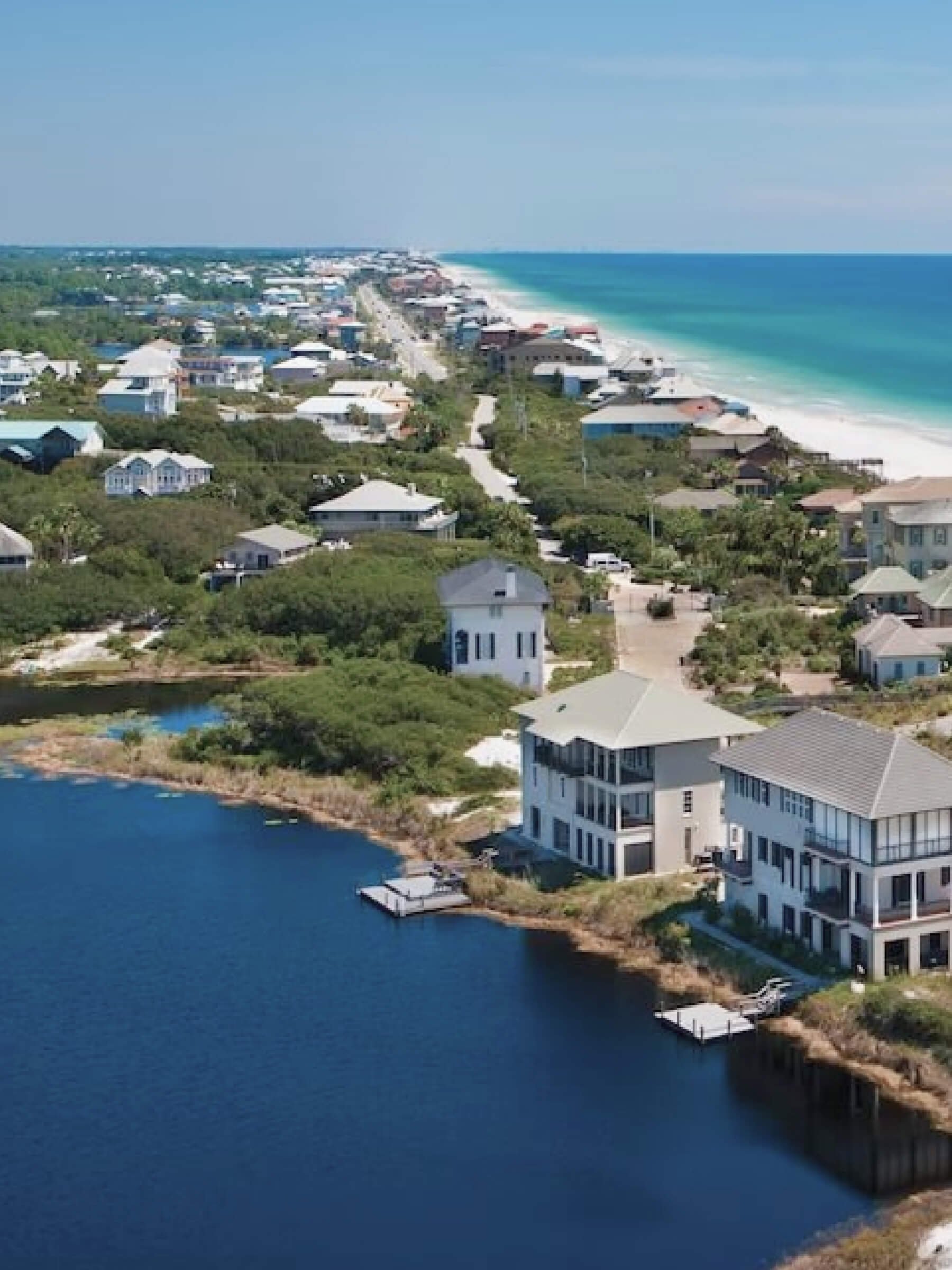 Aerial view of a coastal neighborhood with houses by a calm body of water, with the ocean and a sandy beach in the background.