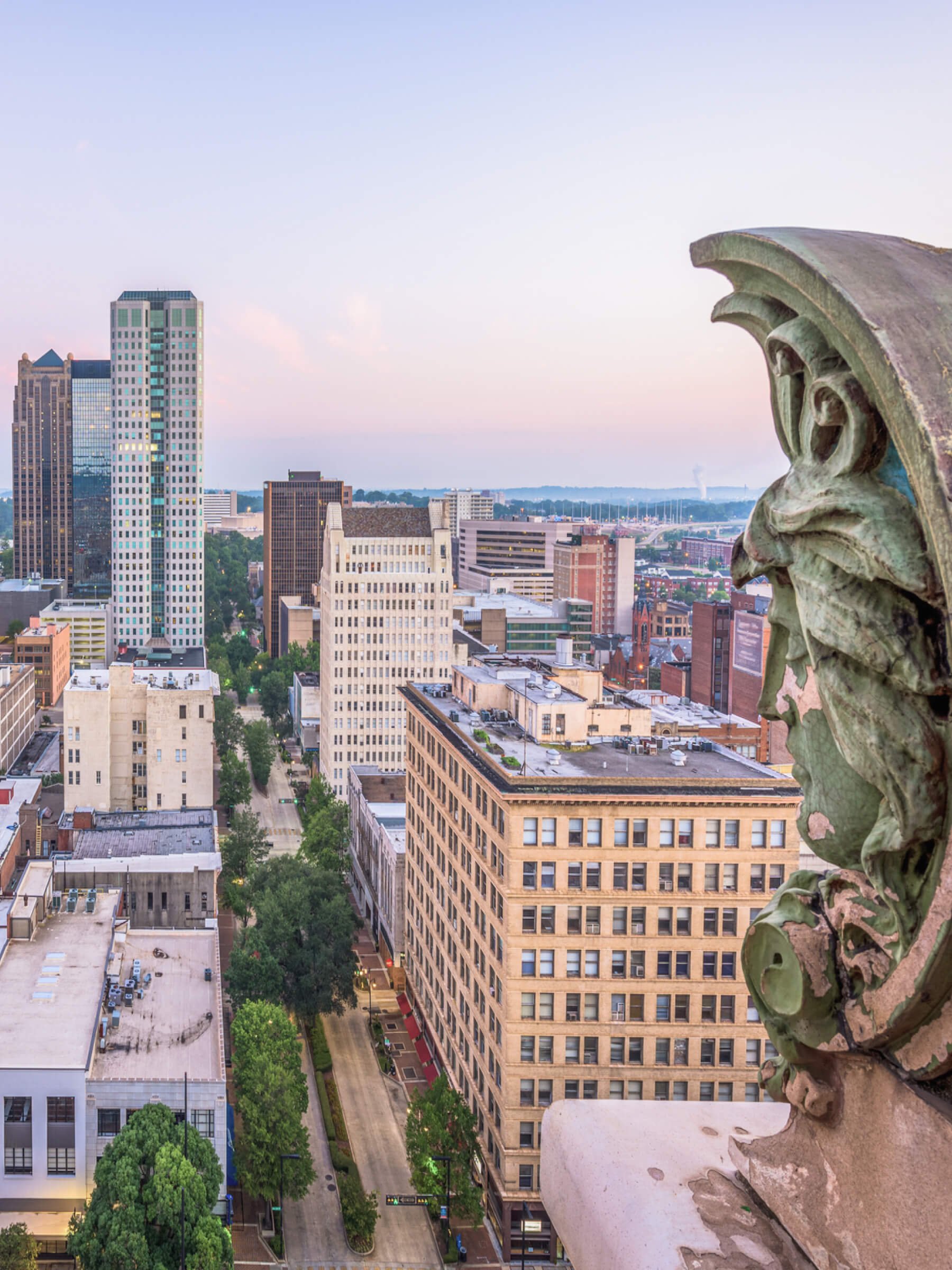 City skyline with tall skyscrapers and historic buildings seen from a high vantage point, with a decorative stone or concrete sculpture in the foreground on the right.