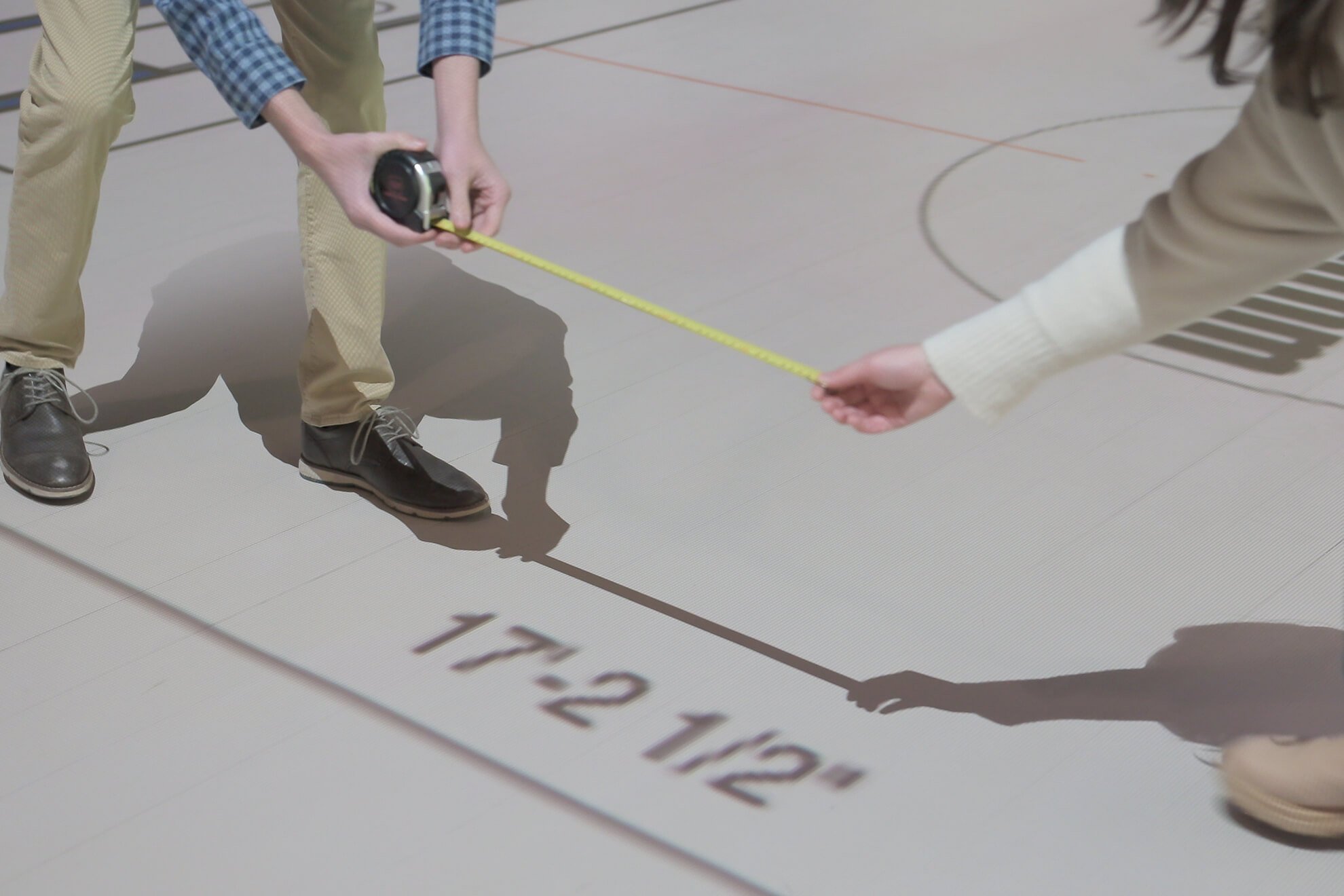 Two people are measuring a line on the floor with a tape measure. One person is holding the tape measure while the other is pulling the tape. The floor has markings and text indicating measurements, including '17.2 ft'.