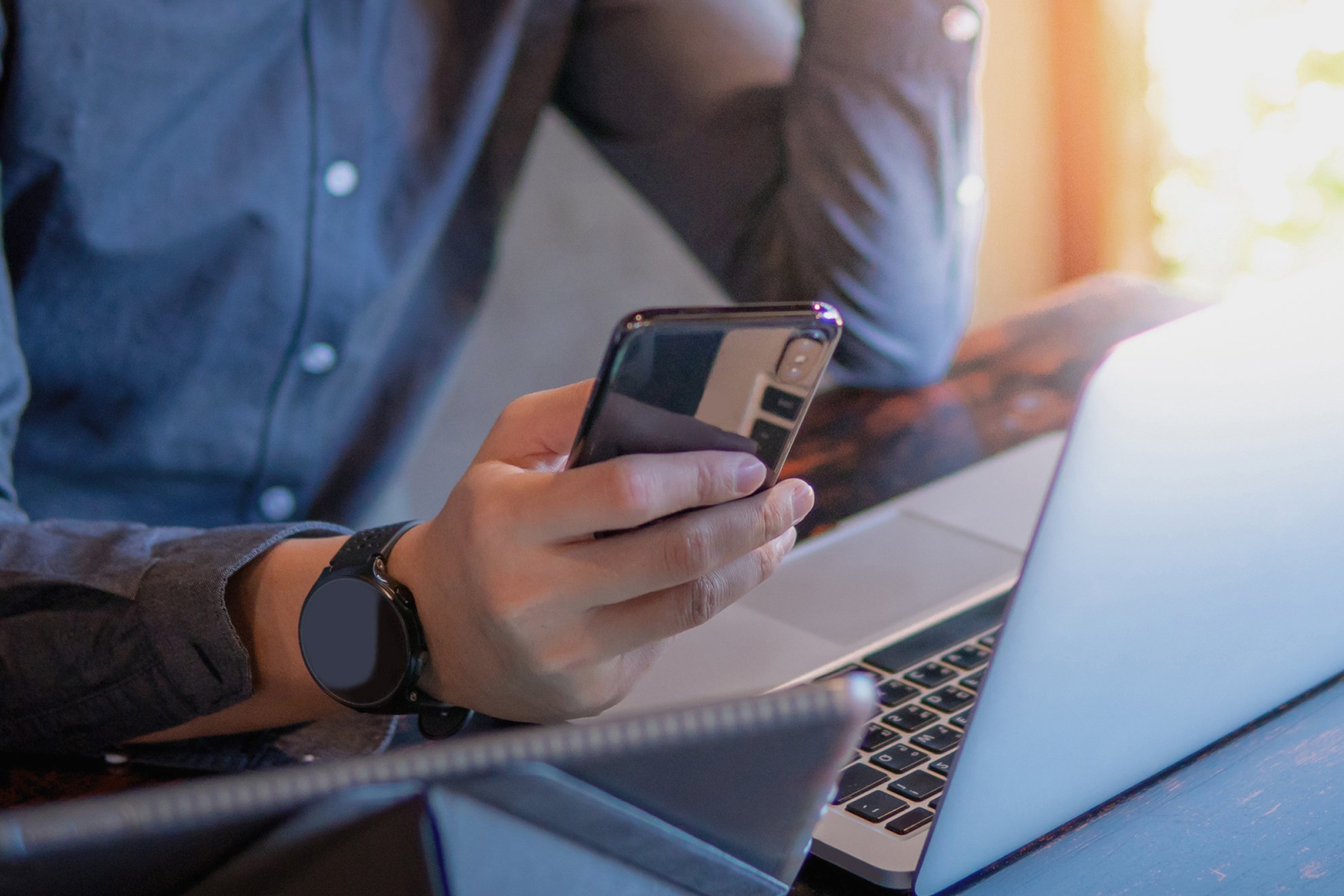 Person wearing a smartwatch and a gray shirt, holding a smartphone while sitting at a desk with a laptop.