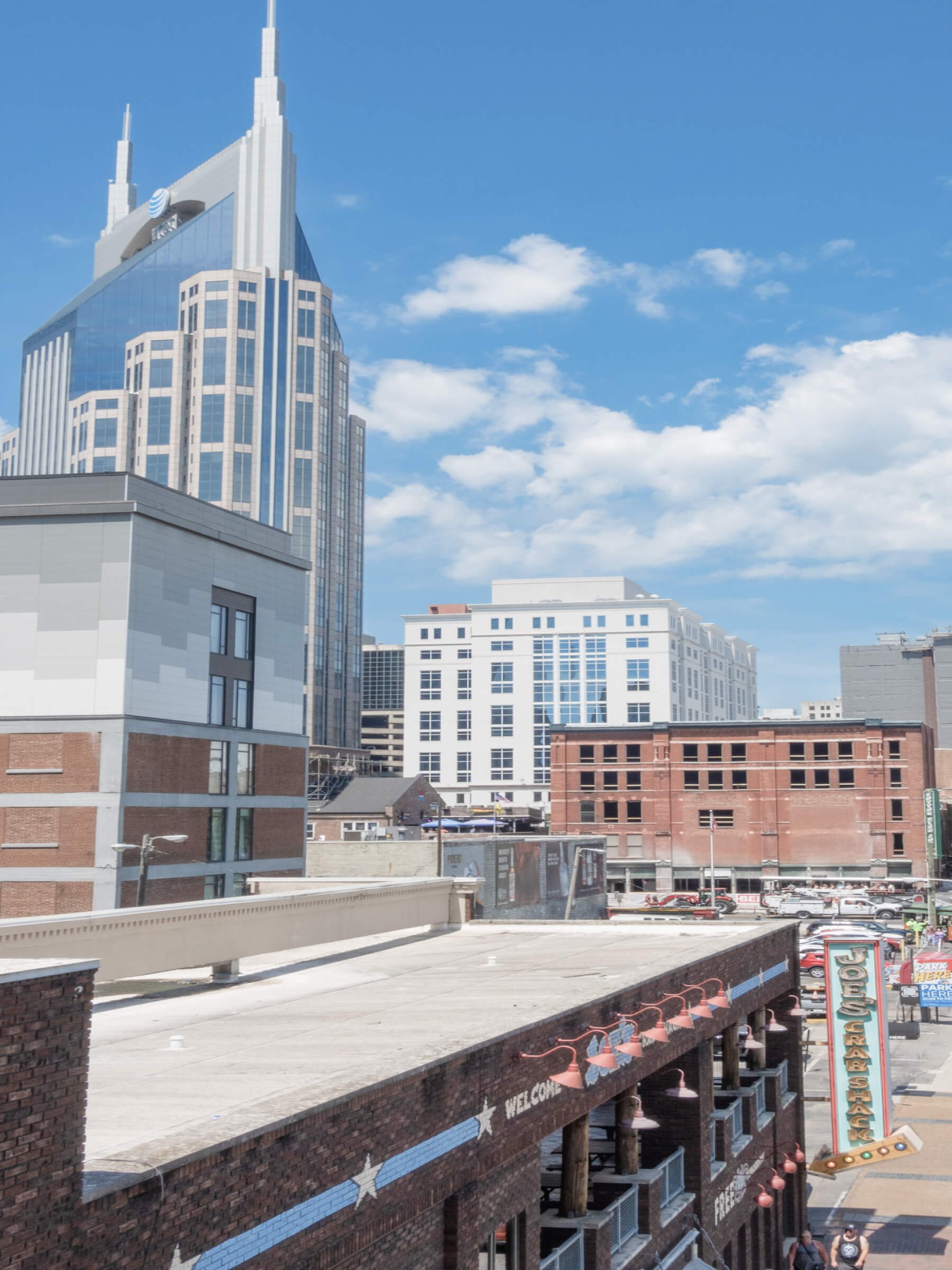 Cityscape featuring modern skyscrapers, a brick building with neon signs, and a partly cloudy blue sky.