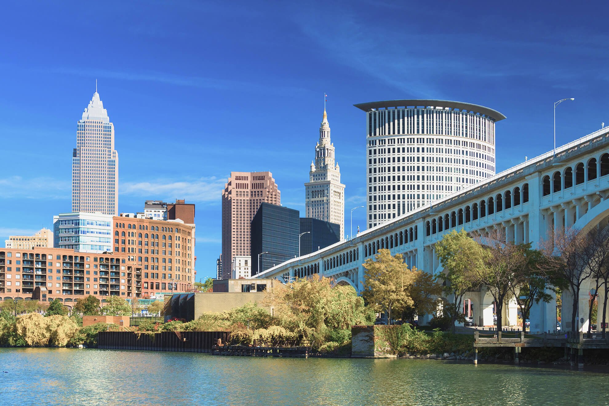 View of downtown Cleveland, Ohio, with tall skyscrapers, a water body in the foreground, and a clear blue sky.