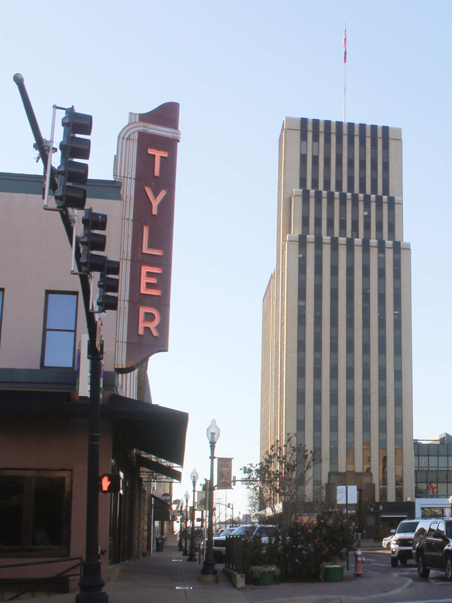 City street scene From Tyler Texas with a vertical marquee sign reading 'TYLER', tall building with flag on top, traffic lights, lamp posts, cars, and storefronts.