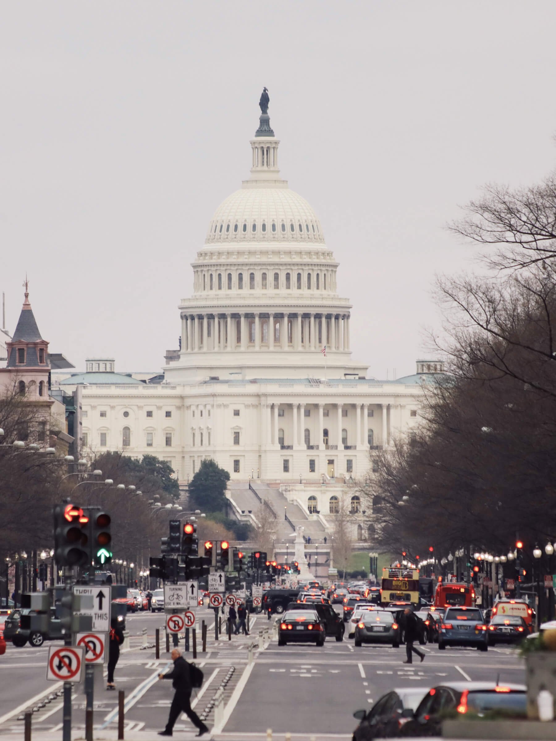 View of the United States Capitol building in Washington, D.C., with cars and pedestrians on the street in the foreground, and leafless trees on the sides.