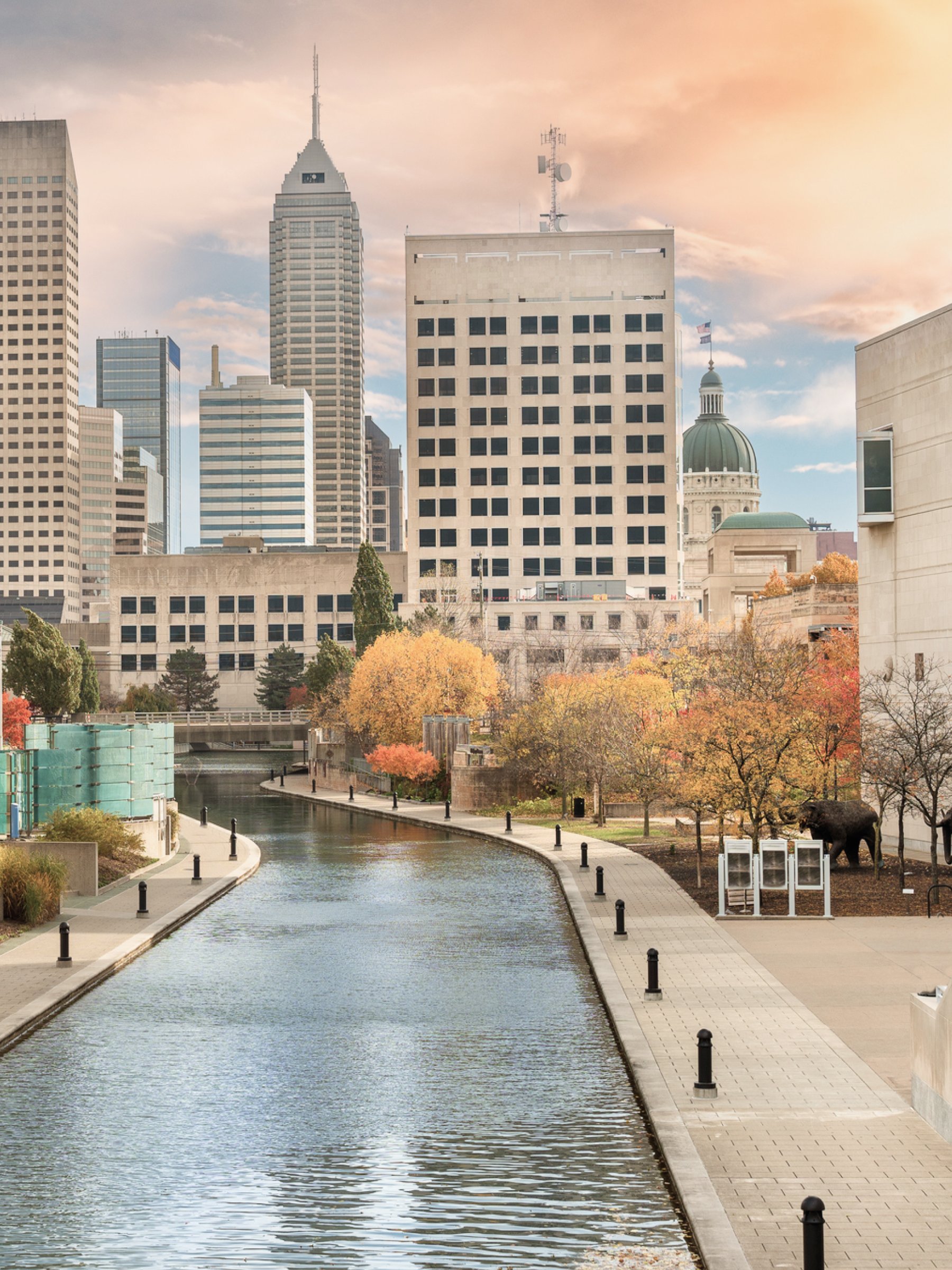 Cityscape with tall modern buildings behind a canal and park with trees in autumn colors.