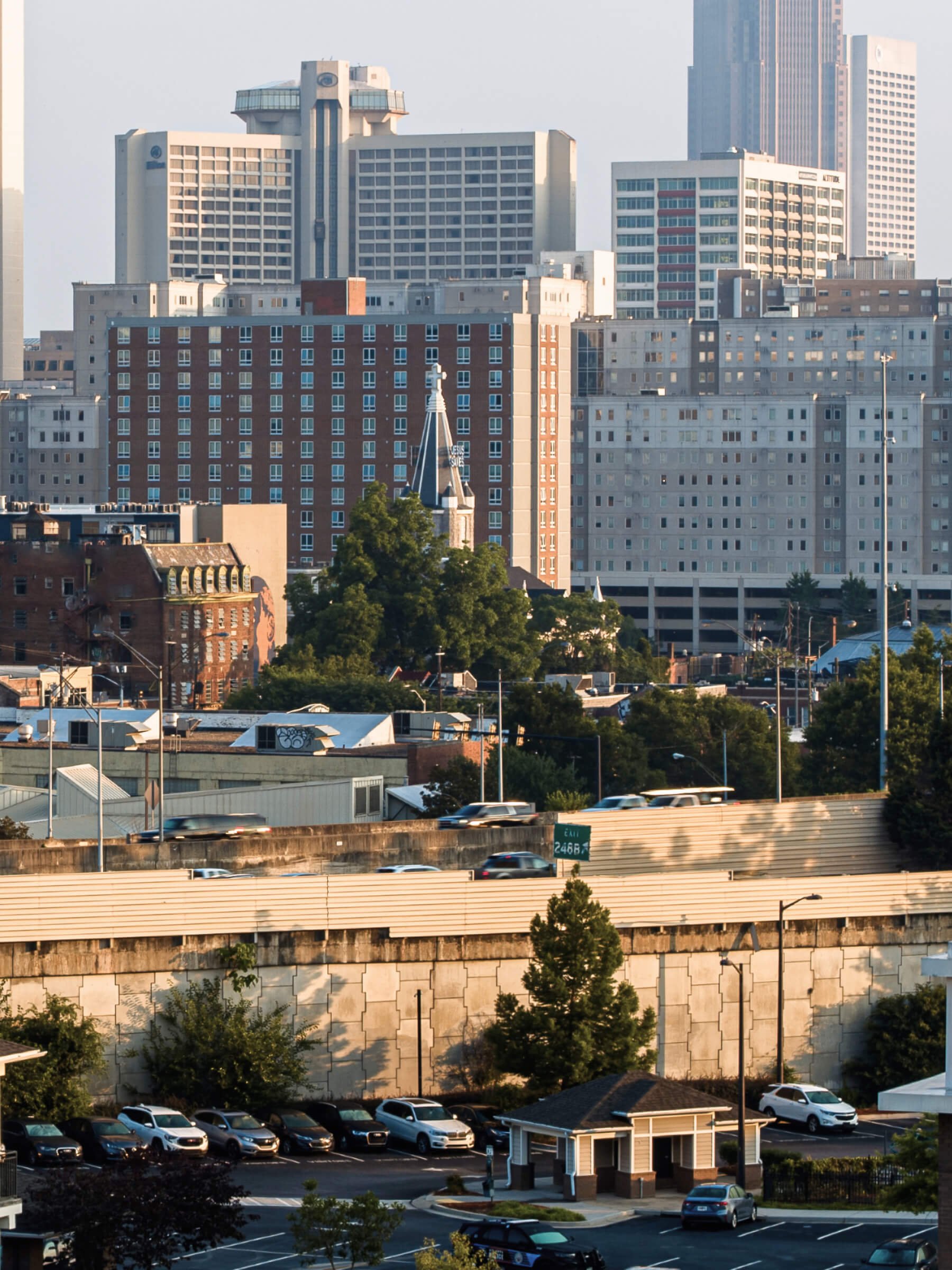 Cityscape with high-rise buildings, a church with a steeple, green trees, and a parking lot with cars.