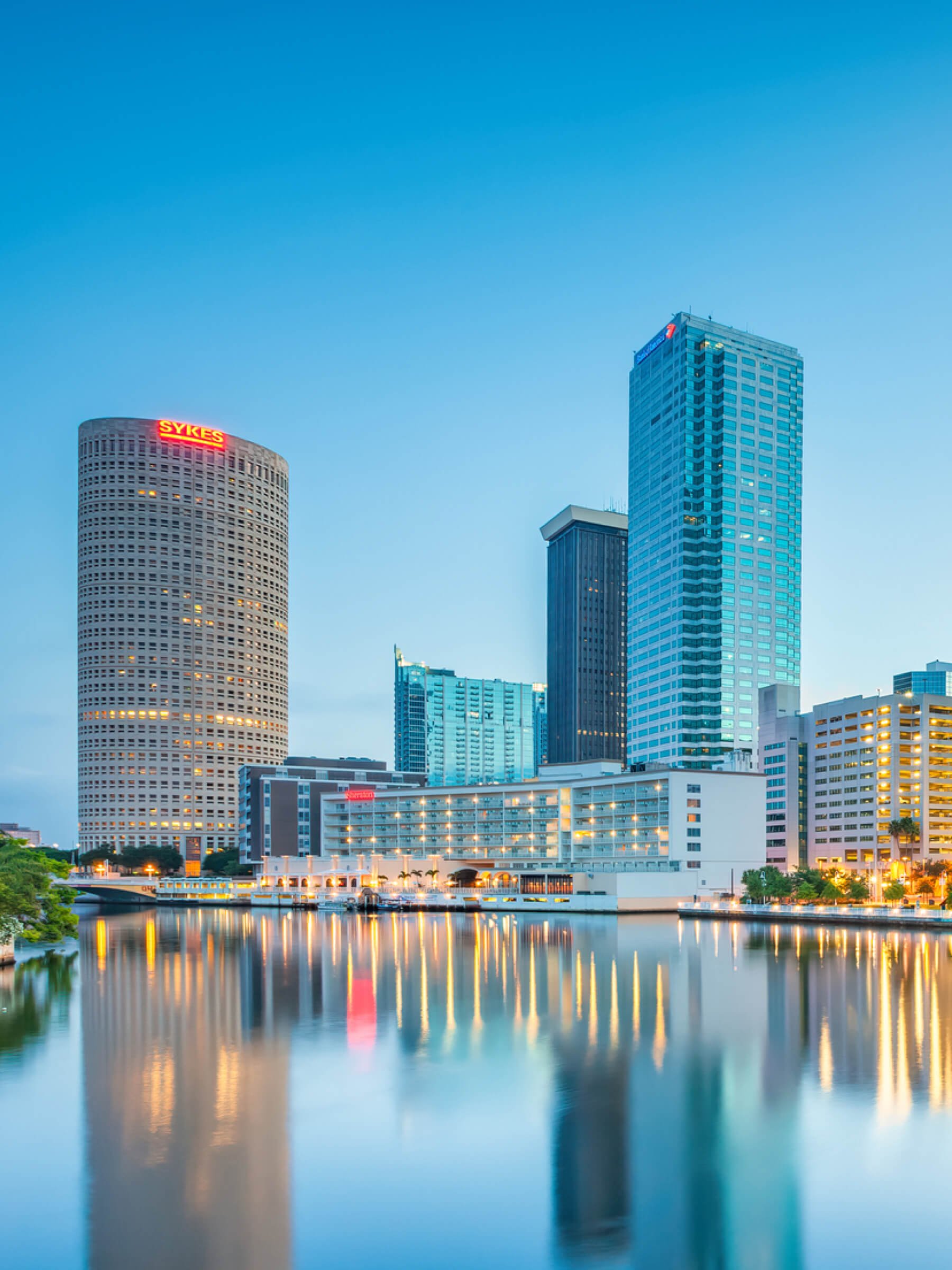 City skyline of Tampa, Florida, with high-rise buildings reflected in water at dusk, with a clear sky and illuminated windows.