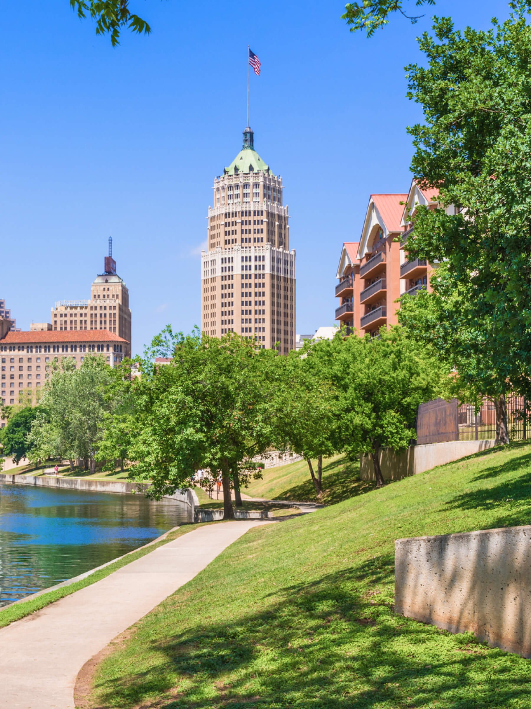 Cityscape of San Antonio Texas with tall buildings, riverwalk, vibrant green park and a clear blue sky.