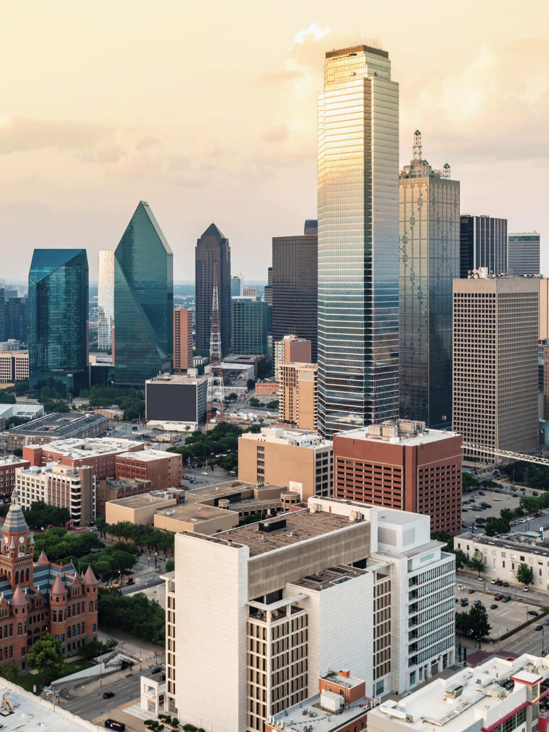 A city skyline featuring tall skyscrapers, including a glass high-rise reflecting the sunset, and other buildings in various architectural styles, with a mix of modern and historic structures, under a partly cloudy sky.